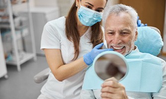 Older man in dental chair looking into mirror with dentist touching his teeth