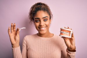 Woman in a brown sweater holding model teeth with braces in one hand and a clear aligner in the other